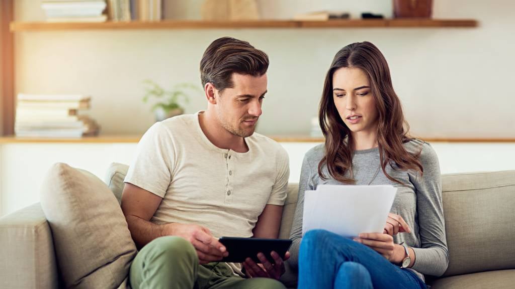 A couple sits on a couch at home reviewing documents together, with one holding a paper and the other using a tablet. 