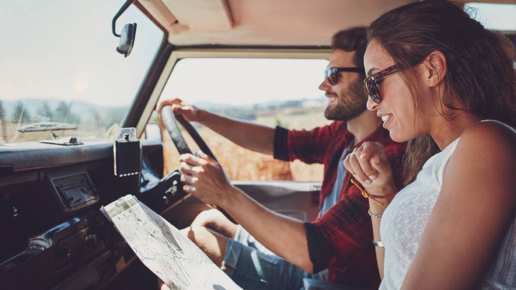 A happy man drives a car and a smiling woman looks at a map next to him