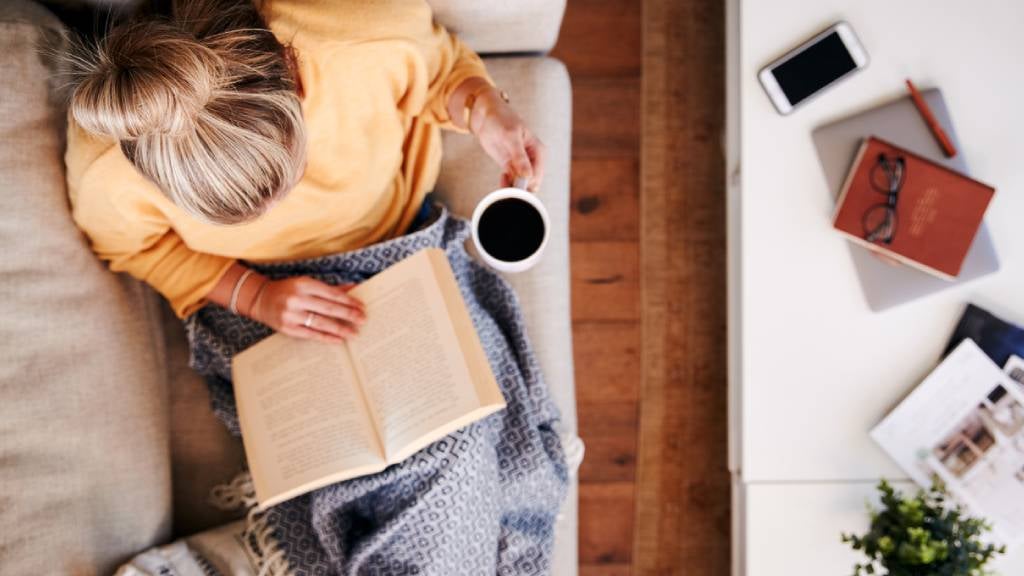 Woman in her thirties reads a book with a black coffee on a couch. 
