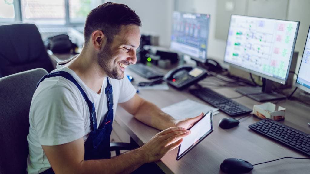 A smiling worker reviews documents on his tablet at his workstation