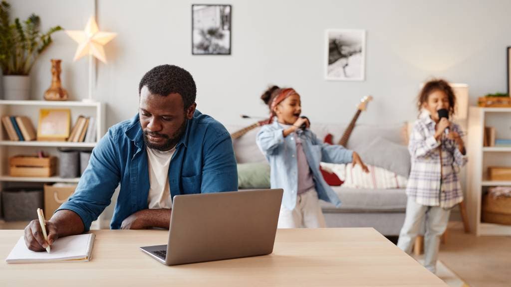 Dad writing at a desk with his daughters singing in the background.