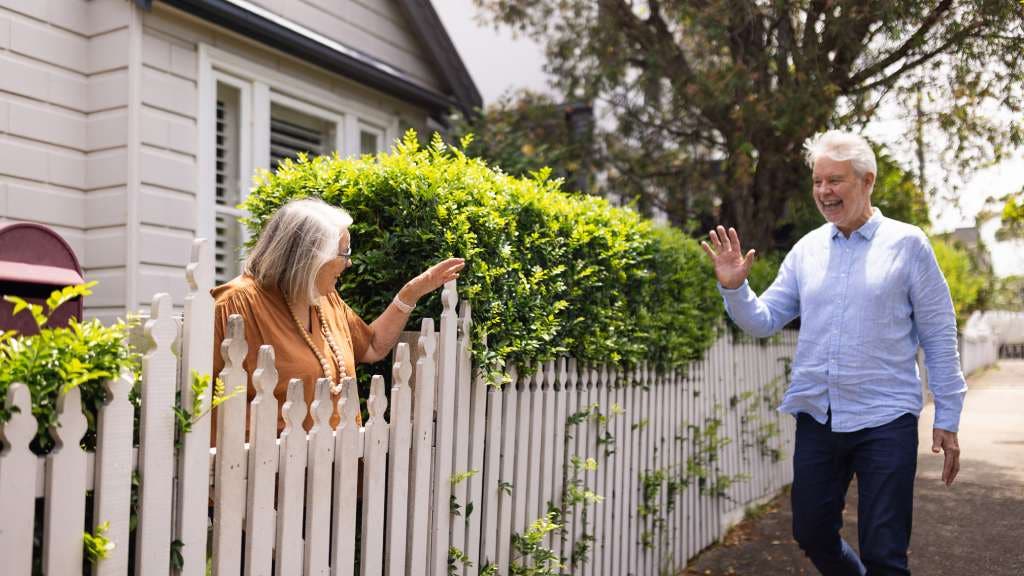 Two cheerful neighbours exchange friendly greetings over a picket fence in a suburban neighbourhood. 