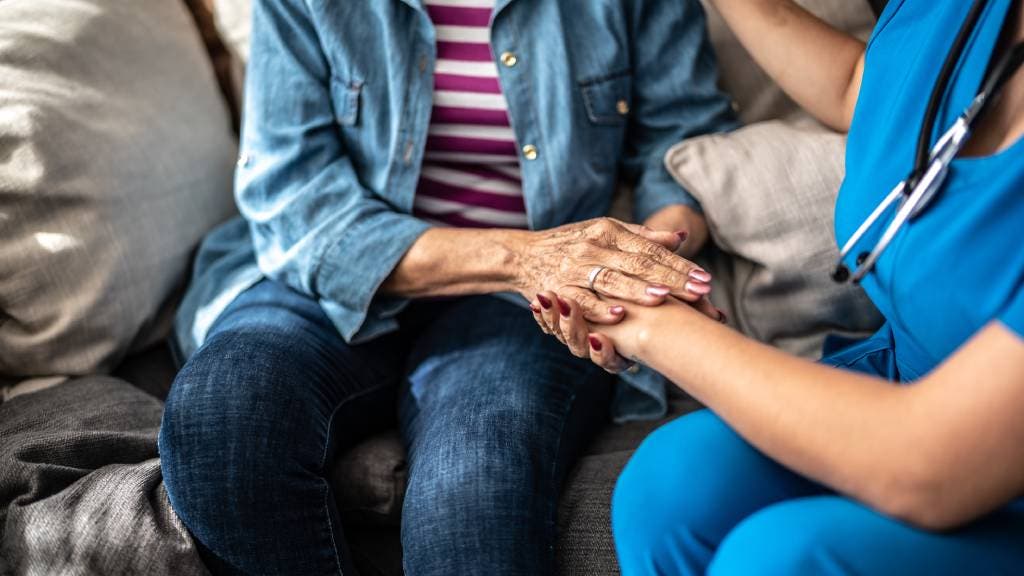 An elderly woman holds the hand of a healthcare worker as they sit on a sofa discussing end-of-life care together.