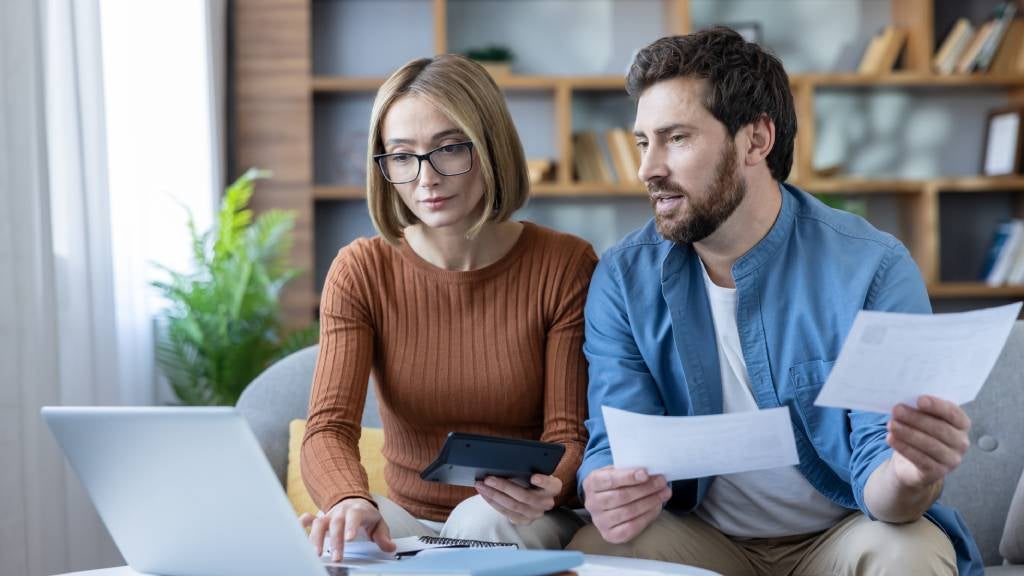 A young couple sit on a sofa in front of a laptop and review financial documents