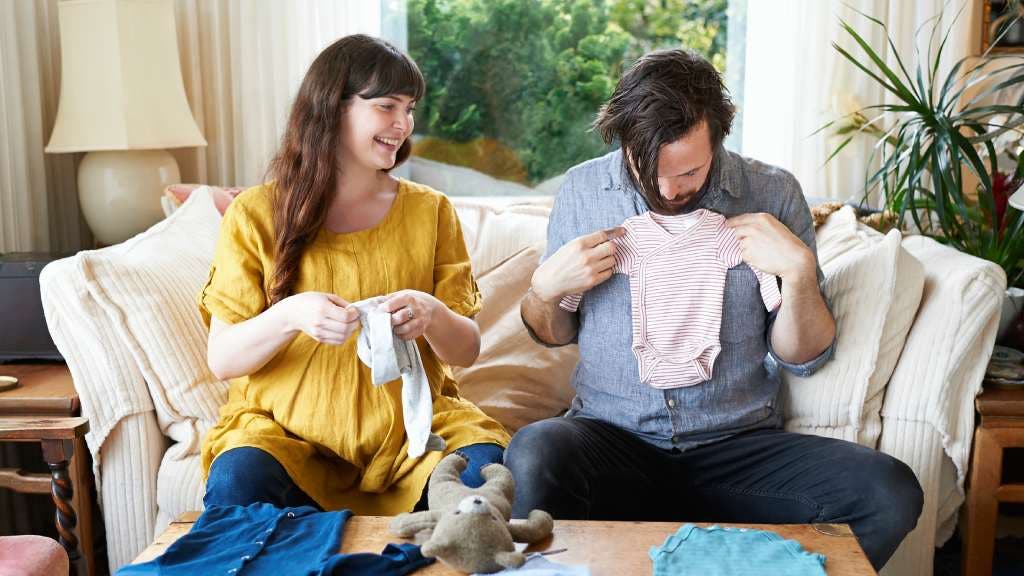 A pregnant woman and her partner sit on a couch, unpacking and playing with new baby clothes.