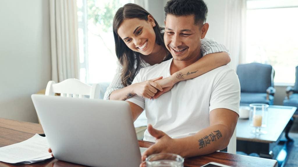 A smiling man looks at a laptop at his desk and a happy woman hugs him