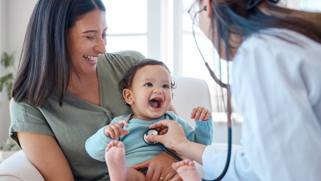 A smiling lady holds her laughing baby who is being examined by a doctor. 