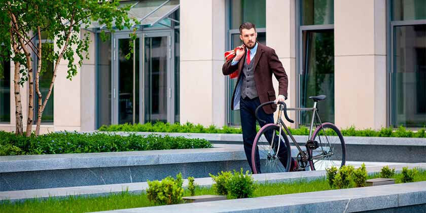 Man in office wear holding a red bag and walking with a bicycle in green office garden 