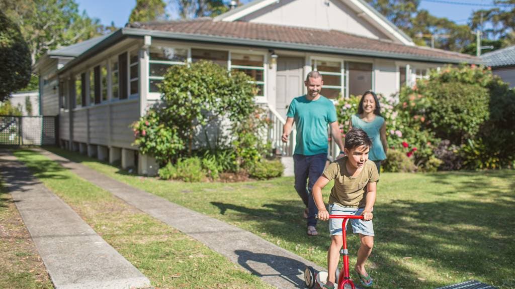 A boy rides his bicycle in his front yard and his smiling parents walk behind him 