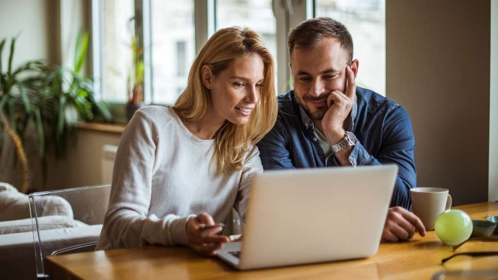 A woman and a man sit at a wooden desk using a laptop together. 