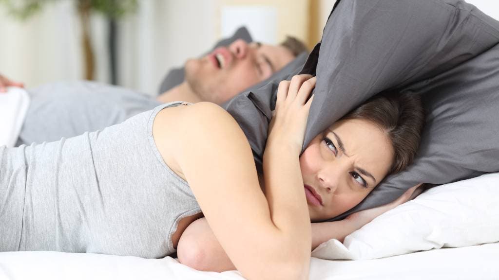 An annoyed woman covers her head with a pillow as a man snores in bed