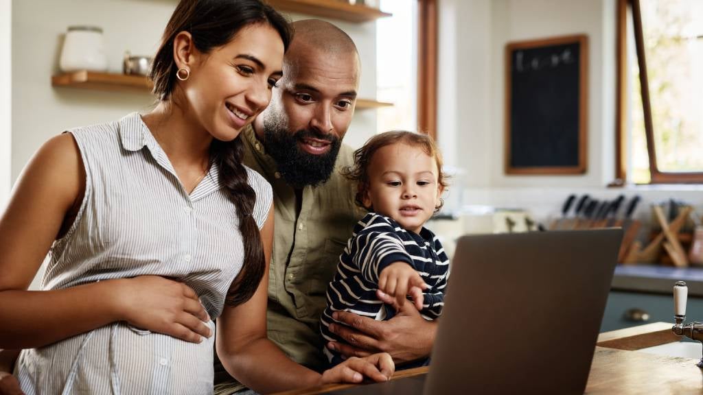 A smiling pregnant woman, her husband and their toddler look at a laptop 
