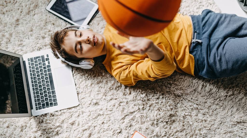 Child laying on floor with laptop open while listening to music and catching a basketball.