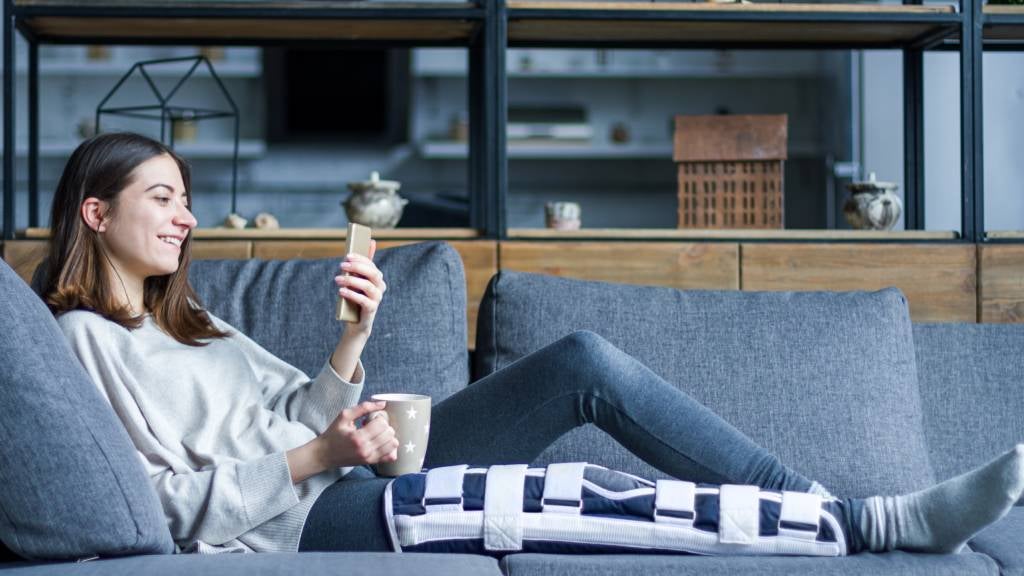 A smiling woman in a leg brace views her phone while sitting on a sofa