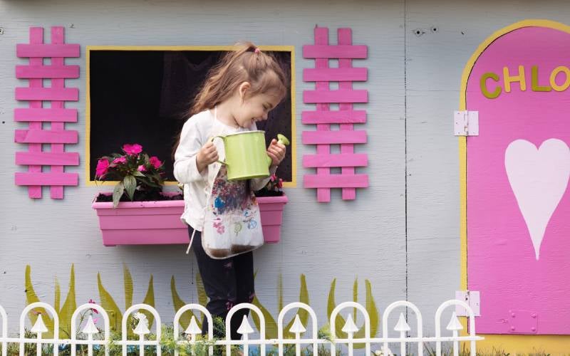 Young girl with watering can in front of cubby house with pink doors and shutters