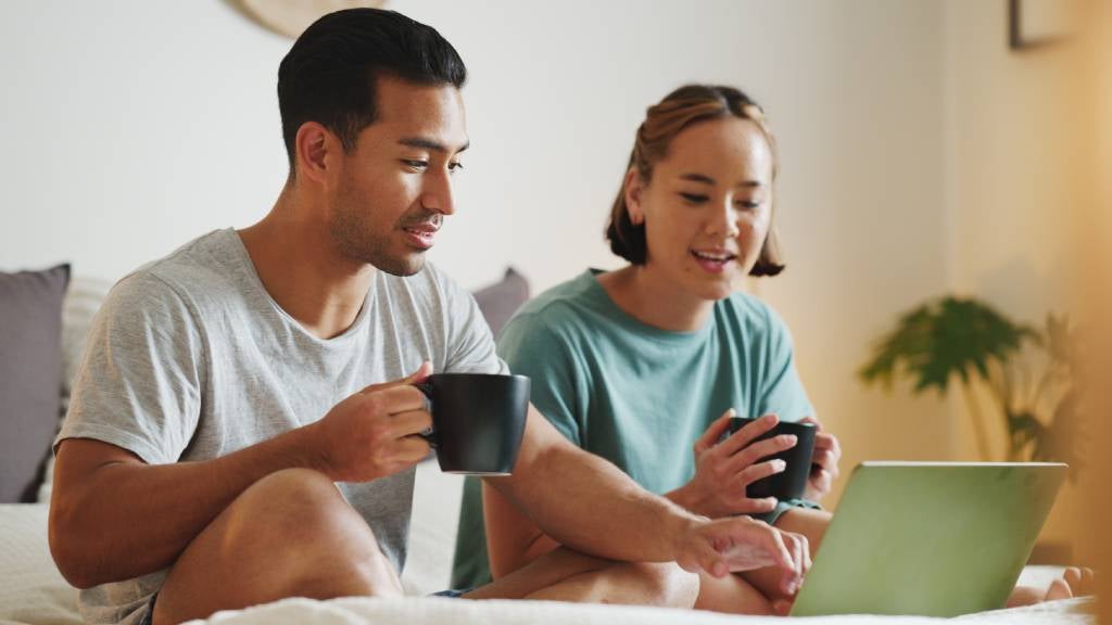 A couple holding coffee cups sit on their bed in front of a laptop