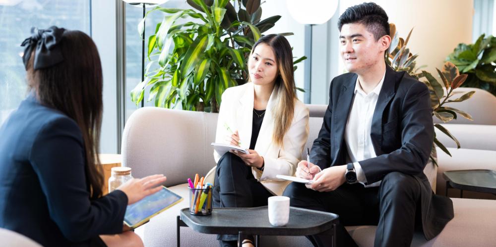 A man and a woman sitting on a business lounge, with pen and paper in hand taking notes, talking to another woman dressed in a dark blue suit.