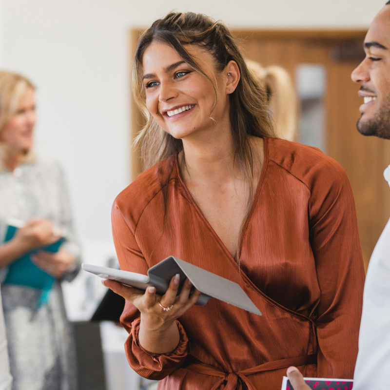 Women with mustard coloured silky shirt and laptop standing near a man and learning forward smiling at someone else