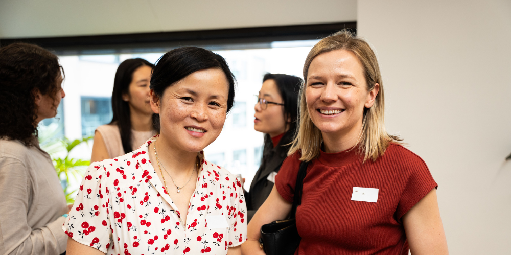 Two females smiling at camera one with dark hair and shirt with red flowers.