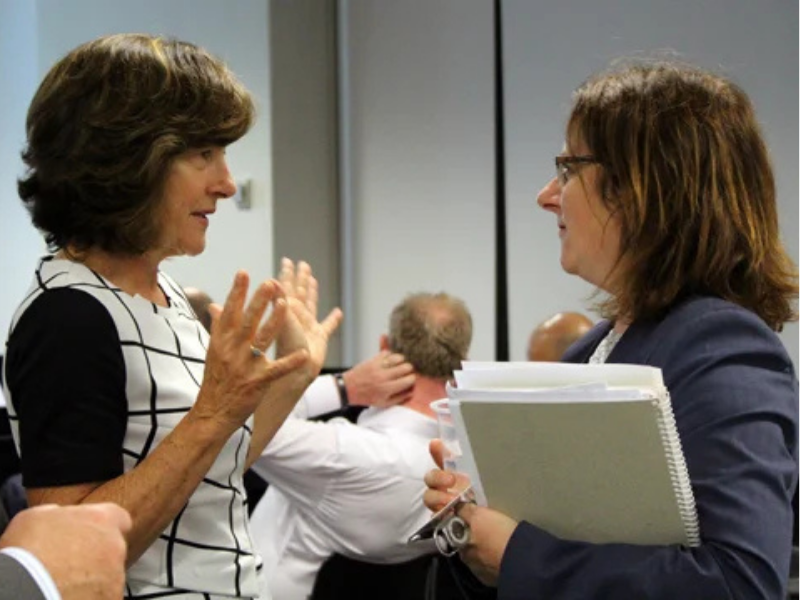 Two women in conversation at a professional event, one gesturing with her hands while holding documents.