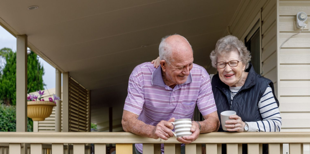 Elderly couple on the porch of their house, enjoying a hot beverage together whilst sharing a laugh