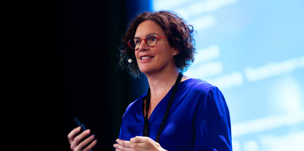 Female presenting with dark curly hair and blue shirt with powerpoint in background