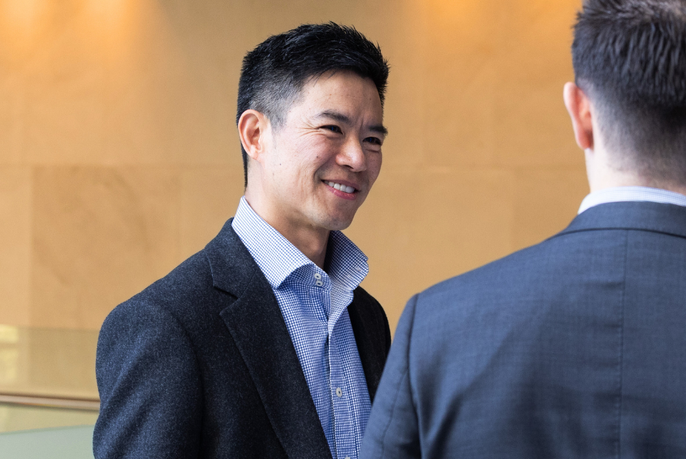 Male dark hair in dark blue suit and open blue shirt smiling and at another male with back to camera