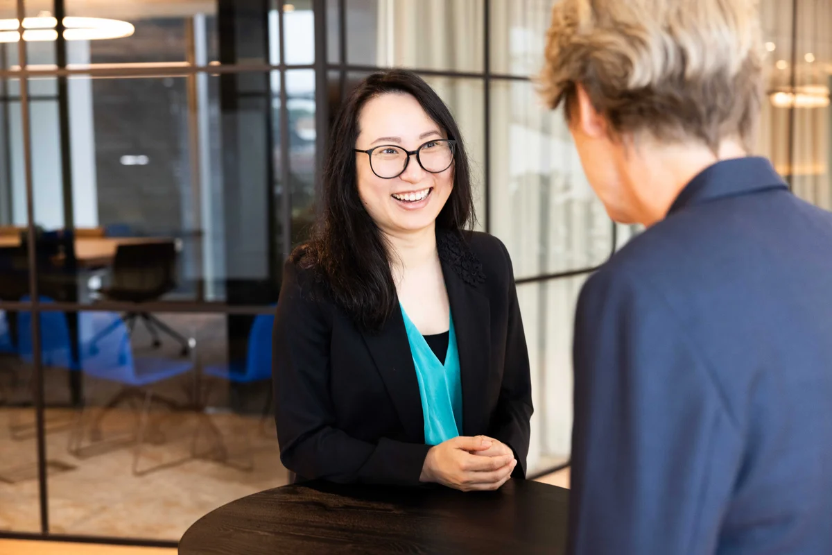 Woman in office in business suit and green blouse, leaning on a high table and talking to a colleague