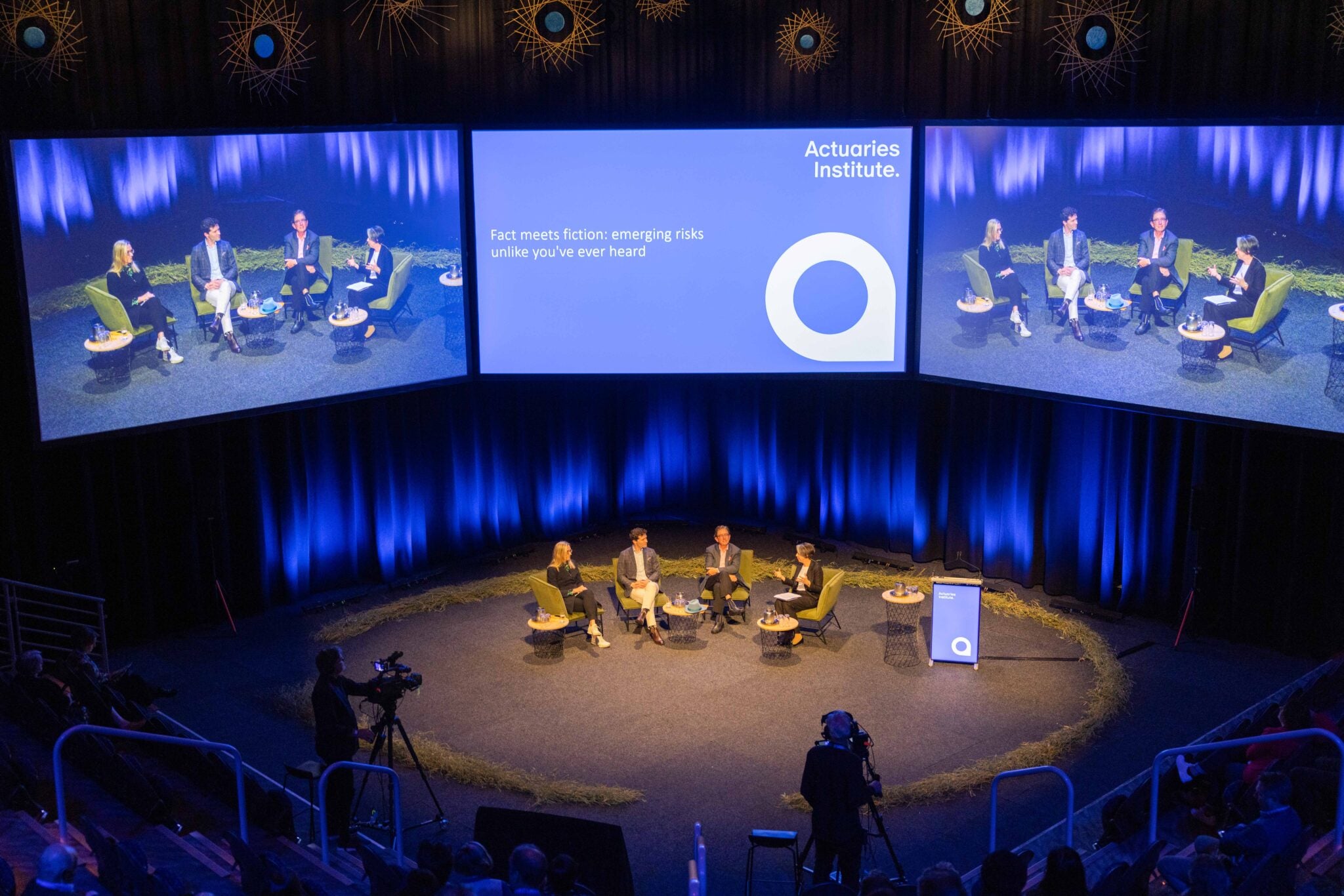 Photo from the All Actuaries Summit Gold Coast, with four presenters in the center stage, and three massive screens above them