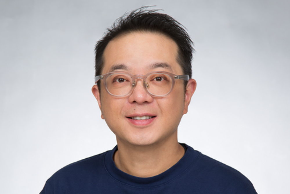 Male with see opaque glasses and dark hair looking at camera. With navy top.