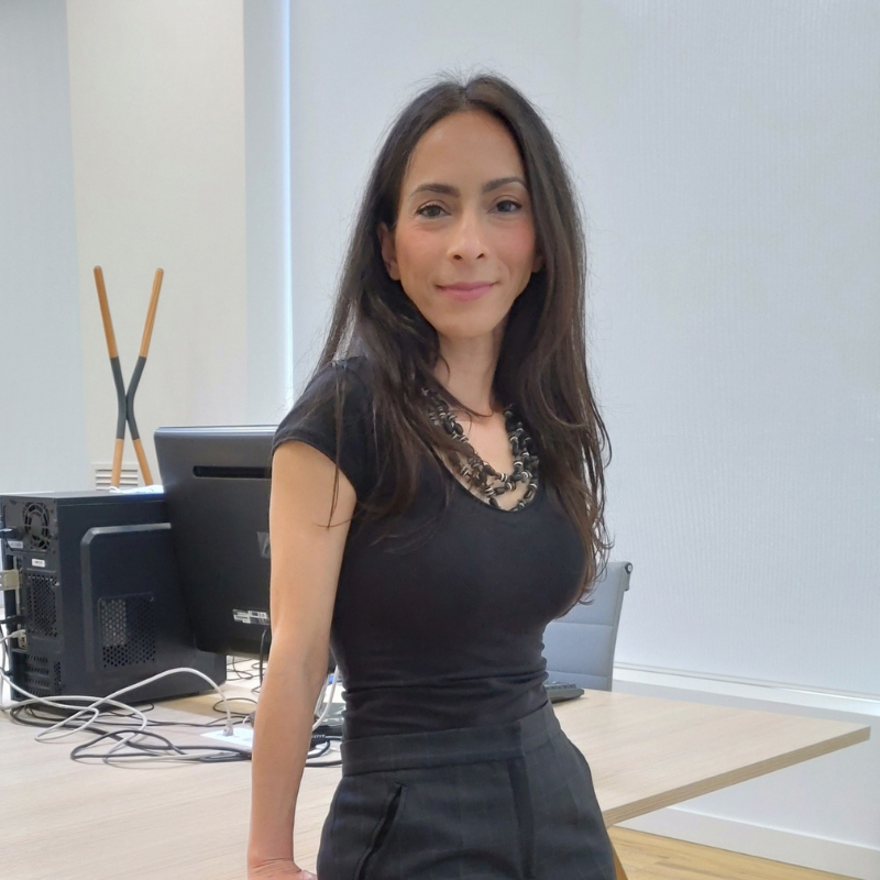 Woman, with long black hair, leaning on desk with black top and trousers