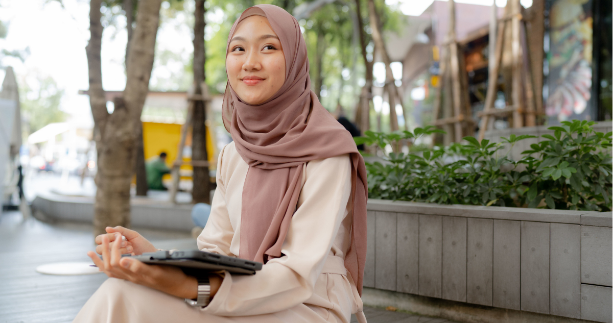 A young woman wearing a hijab sits outdoors at a café, smiling and holding a tablet, with trees and greenery in the background.