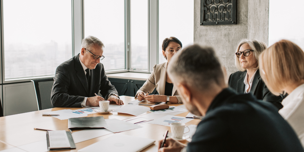 Men and women in business attire sit at a boardroom table.