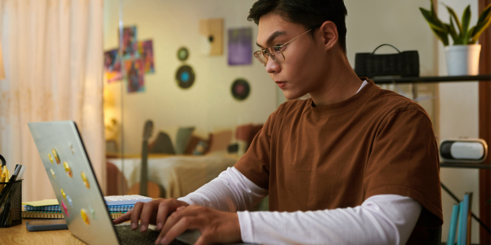A young man wearing glasses typing on a laptop at a desk in a home study space