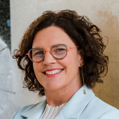 Business women with light blue jacket, short brown curly hair and orange speckled glasses smiling to camera.