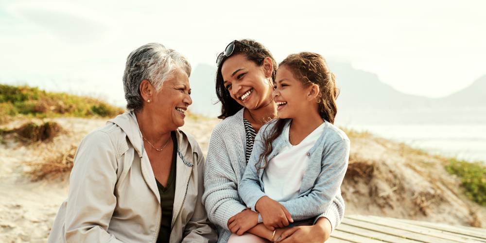 Three generations of women — a grandmother, mother and young girl — sit together on a beach boardwalk, smiling and laughing in warm afternoon light.