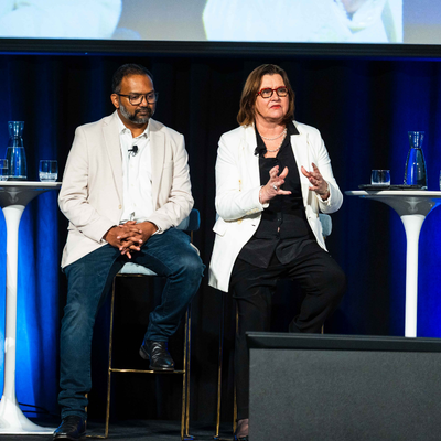Jacki Johnson speaking with hands raised while Ian Hamm and Suhit Anantula listen during the opening plenary