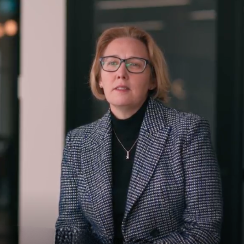 female in black and white checked jacket in office talking to camera with short blond hair and black glasses