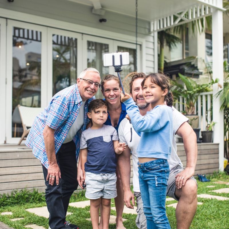 A three-generation family of five — a grandfather, two parents and two young children — pose together for a selfie on a stick outside a light-coloured weatherboard home. All are smiling and casually dressed. Lush green plants are visible in the background.