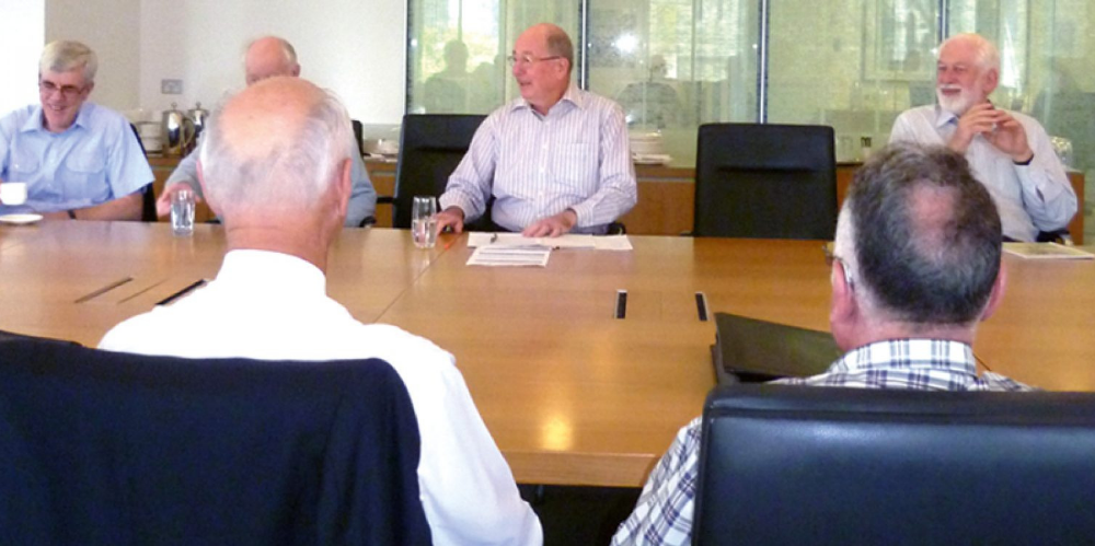 A group of older men seated around a boardroom table in relaxed discussion at a professional meeting.