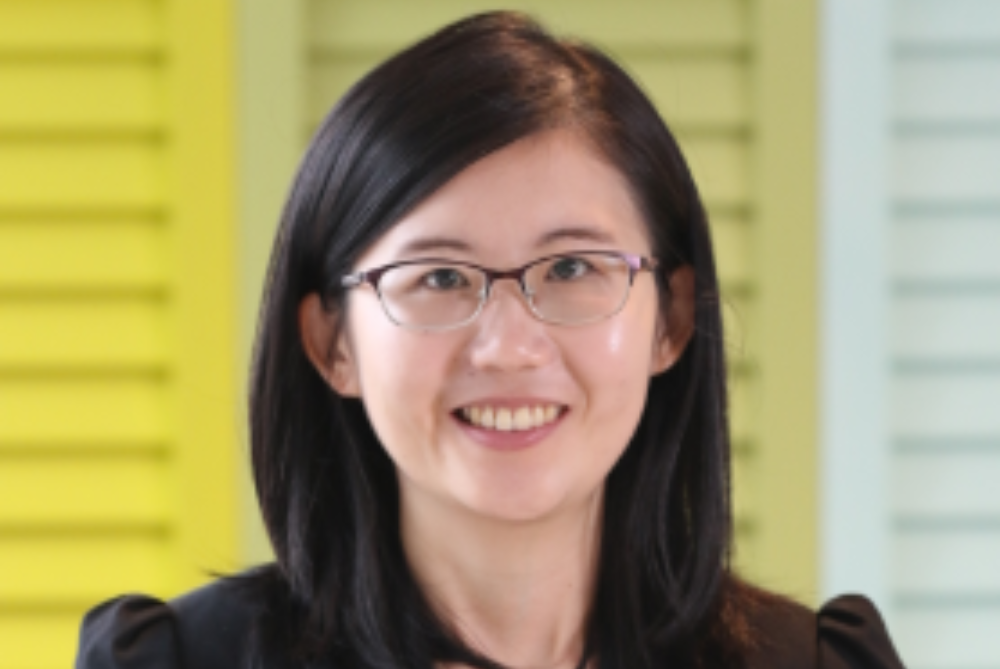 Woman smiling at camera, with black shoulder length, hair, black glasses with a bright yellow background.
