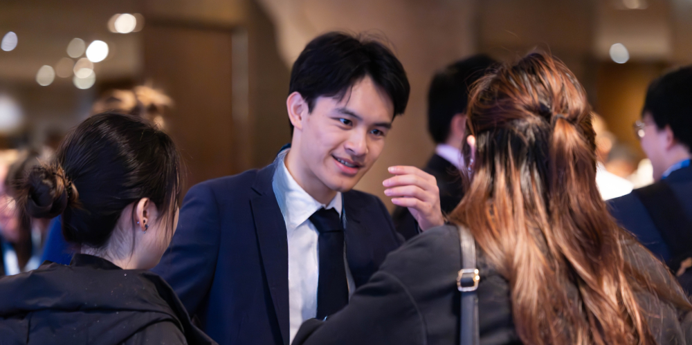 Man in suit with dark hair leaning forward with a hand raised smiling and talking to two females