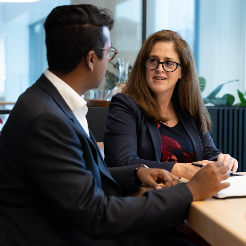 Man on left in suit and business woman on right sitting at a table with papers and pens, talking to eachother