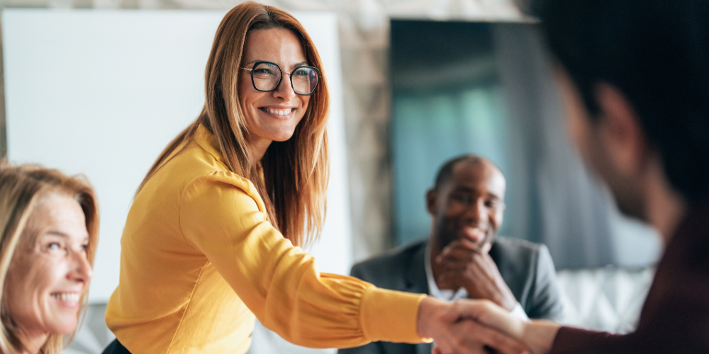 A smiling businesswoman in a yellow blouse and glasses confidently shakes hands with a colleague during a corporate meeting