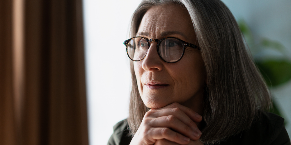 Thoughtful mature woman with glasses looking away, contemplating near the window at home or office.