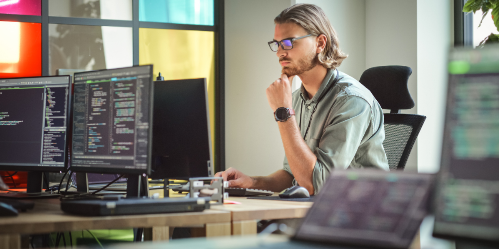 Male with blonde hair and short beard, khaki shirt looking at  the computer, thinking