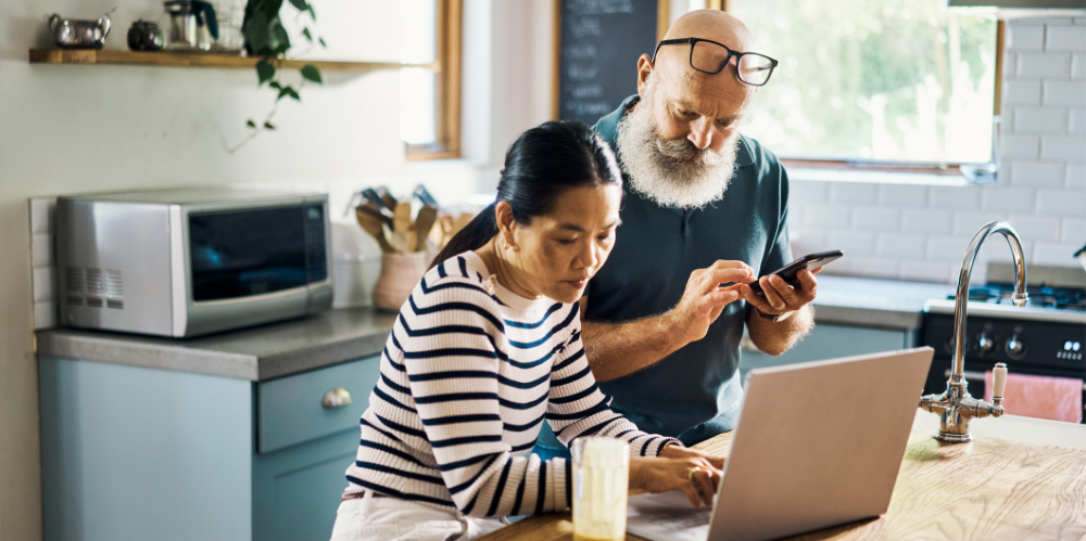 A mature couple using a laptop and phone in the kitchen at home.
