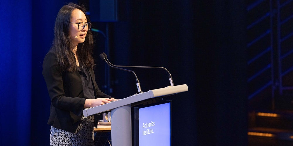 Jess Chen wearing black blazer on stage speaking at Actuaries Institute event.