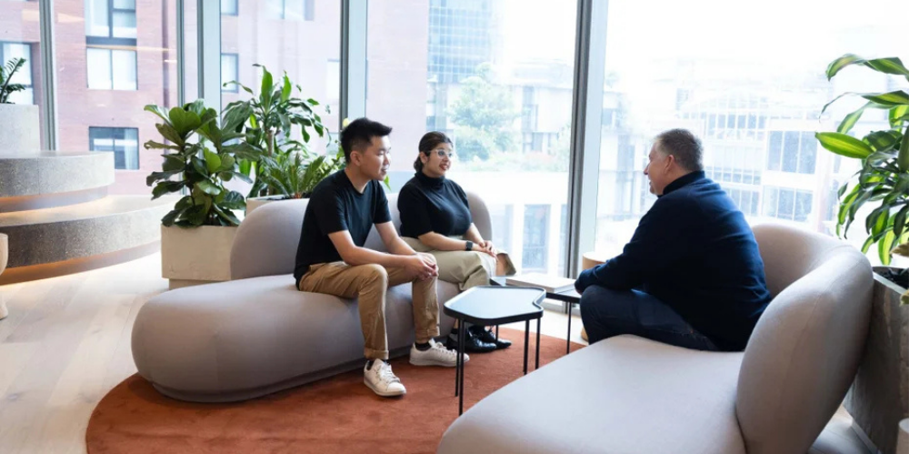 Three professionals in a relaxed conversation seated in a modern office lounge with floor-to-ceiling windows.