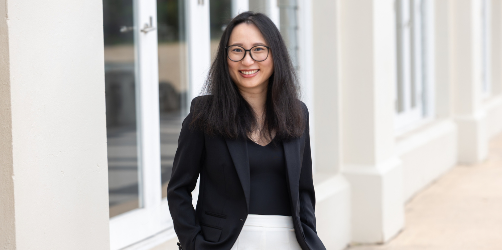 Female with long black hair, black jacket and top with white trousers, smiling and looking to camera and standing in front of white and cream building.